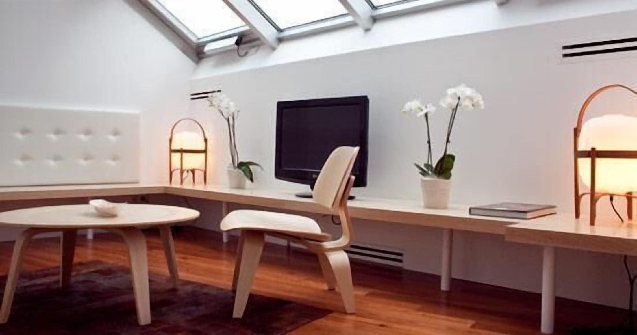 Une pièce moderne et bien éclairée, dotée d’une lucarne, d’un bureau, d’une chaise, d’un téléviseur et de deux plantes en pot.