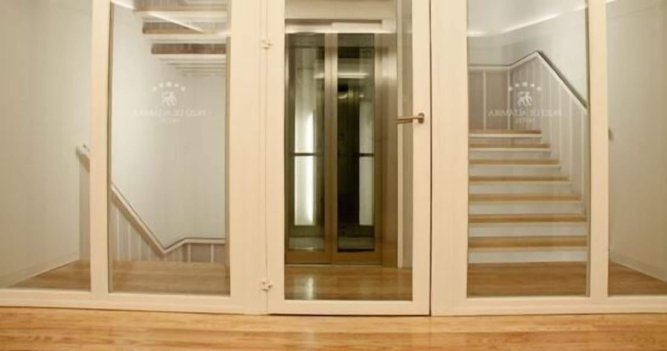 Entrada de un hotel con puerta de cristal, escalera de madera y un logotipo visible en la pared.