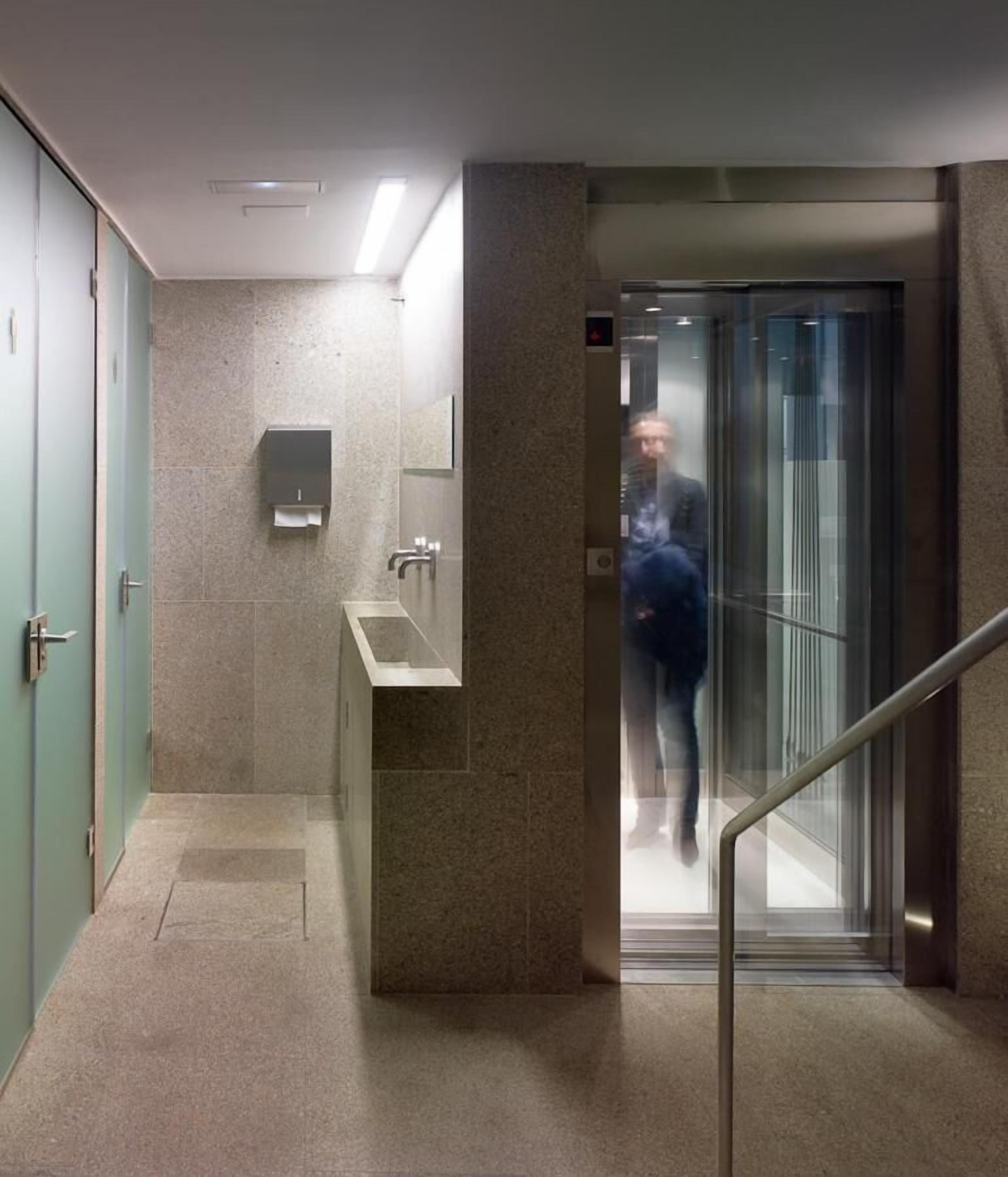 A modern elevator with a glass door and metal handrail, leading to a hallway with a marble floor and a wall-mounted hand dryer.