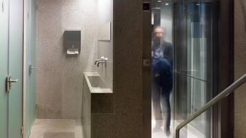 A modern elevator with a glass door and metal handrail, leading to a hallway with a marble floor and a wall-mounted hand dryer.