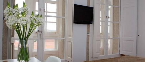 A hotel room with a bed, a TV mounted on the wall, a table with a vase of flowers, and large windows.
