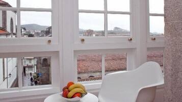 Una habitación con un ventanal grande, una silla blanca y un frutero sobre una mesa.