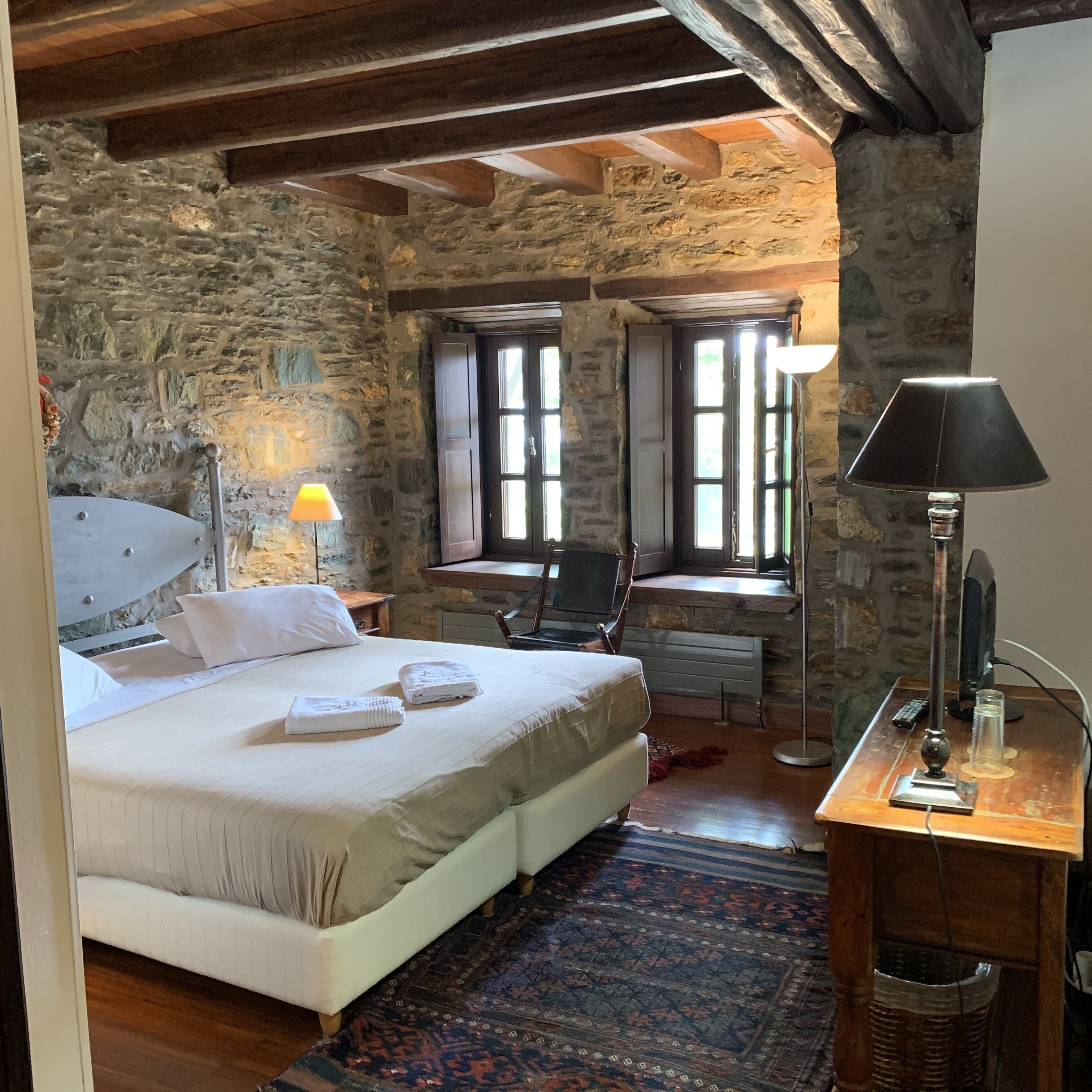 Une chambre avec un mur en pierre, un lit avec du linge de lit blanc, un bureau avec une lampe et une chaise.