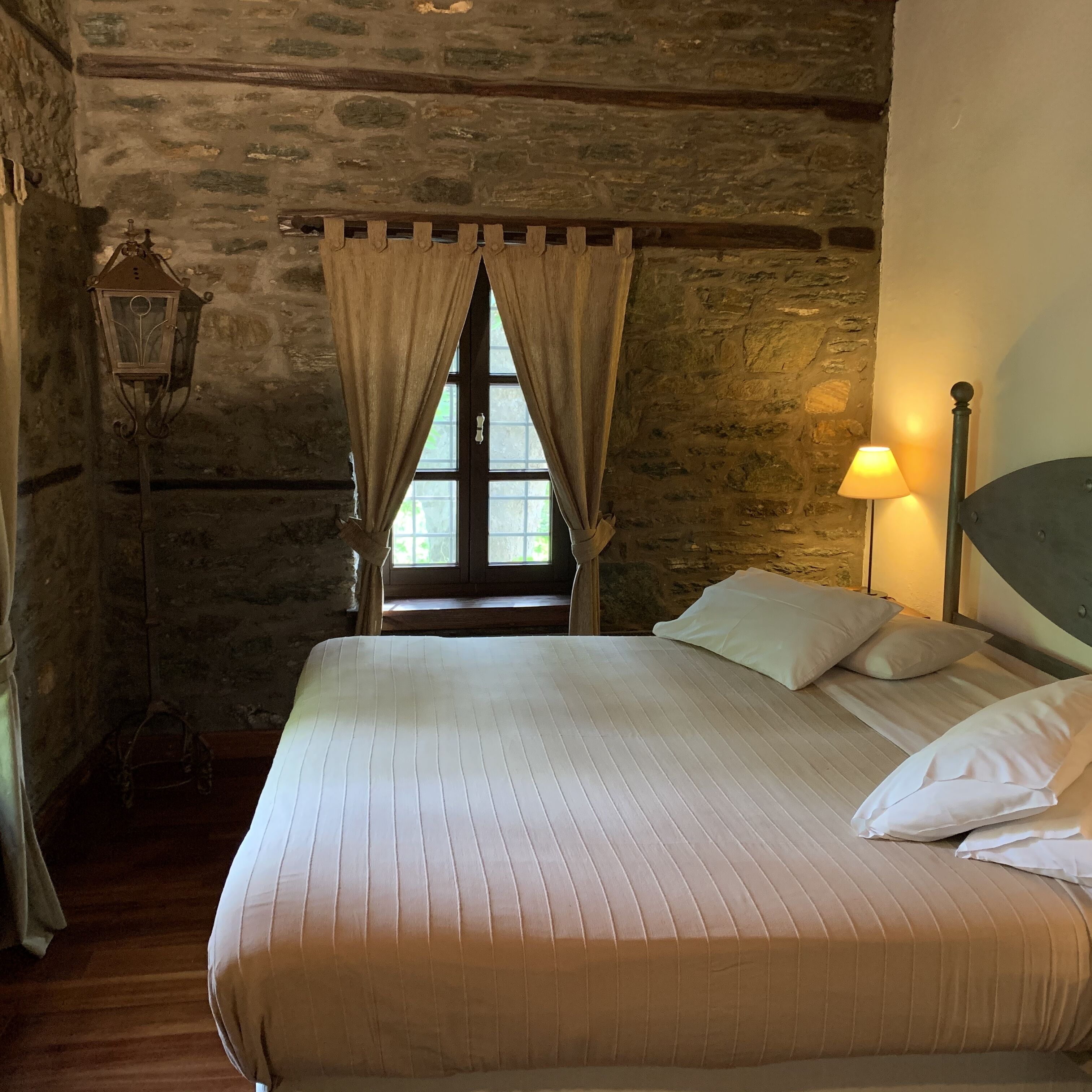 Una camera da letto con parete in pietra, struttura del letto in legno, finestra con tende, abat-jour e lampadario.