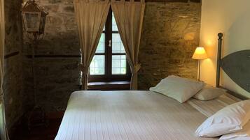 Un dormitorio con pared de piedra, estructura de cama de madera, ventana con cortinas, lámpara de noche y lámpara colgante.