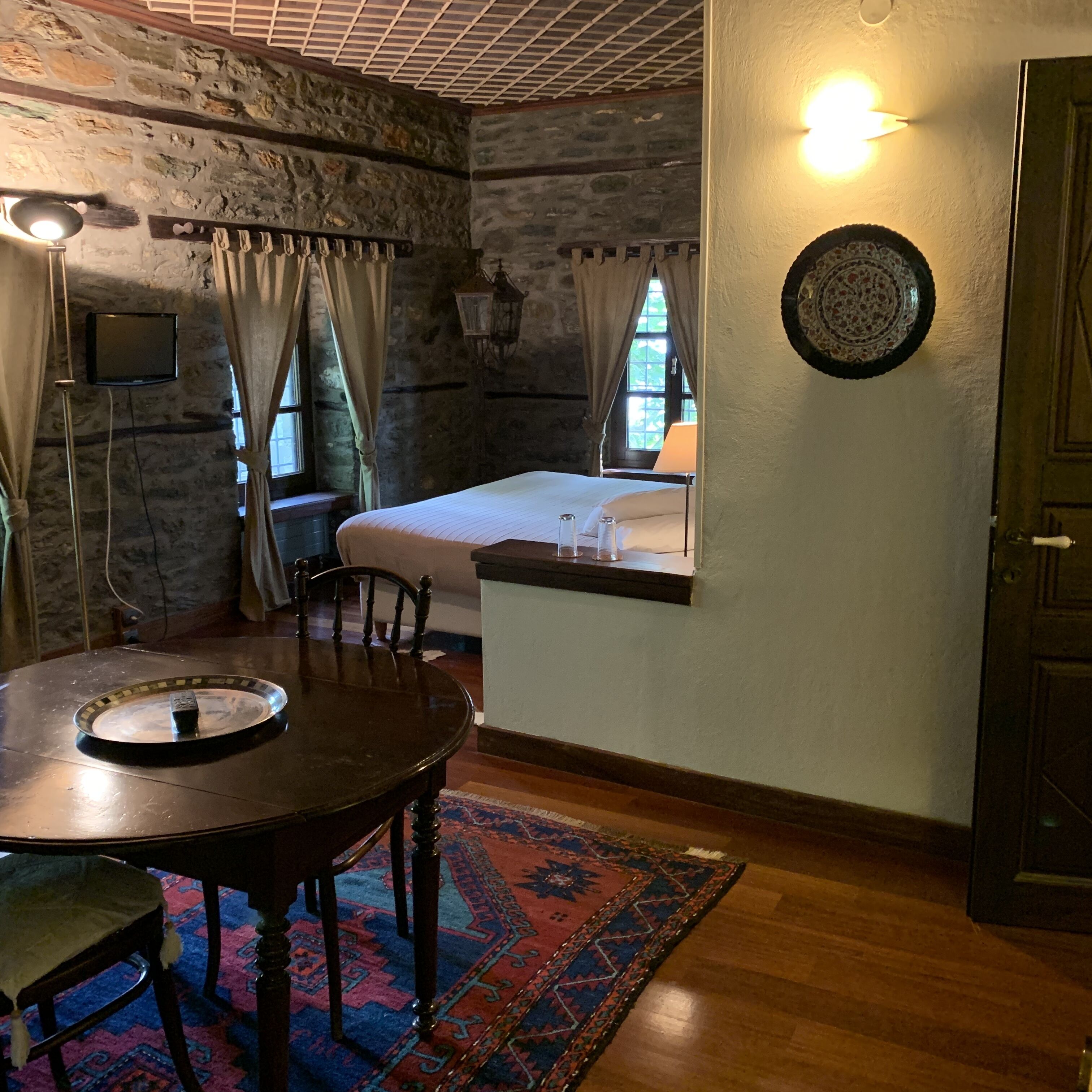 A room with a bed, a table with chairs, a rug, a wall-mounted TV, and a window with curtains.