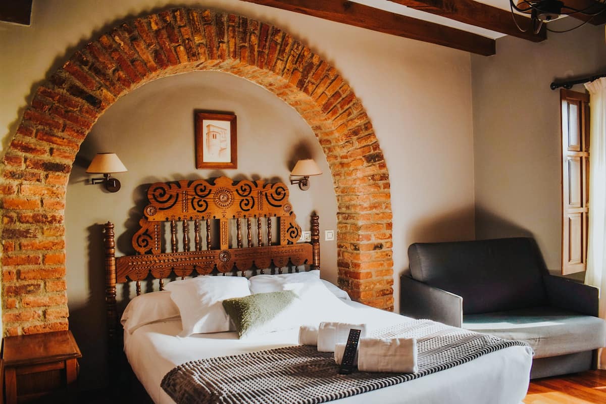 Quarto com cabeceira em arco de tijolos, cama com roupa de cama branca, sofá preto e duas luminárias fixadas na parede.