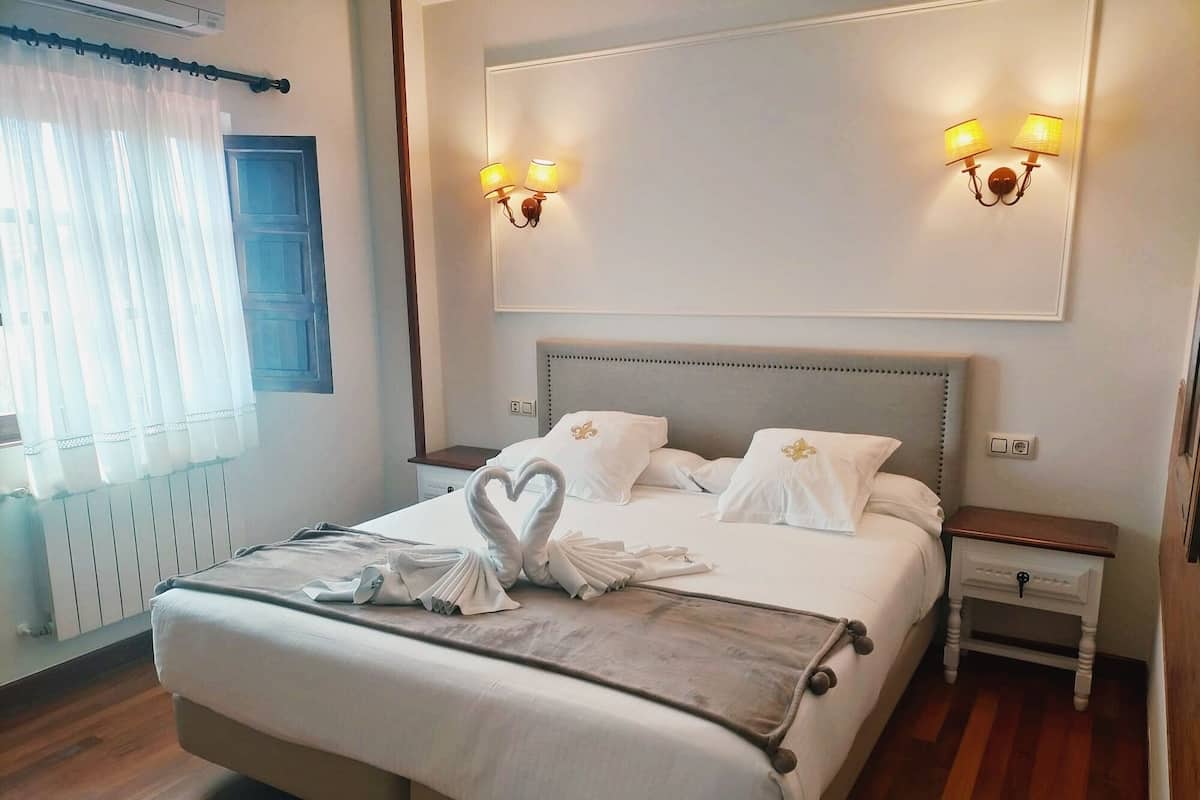 Quarto de hotel com uma cama grande, duas mesas de cabeceira, luminária fixada na parede e uma janela com cortinas.