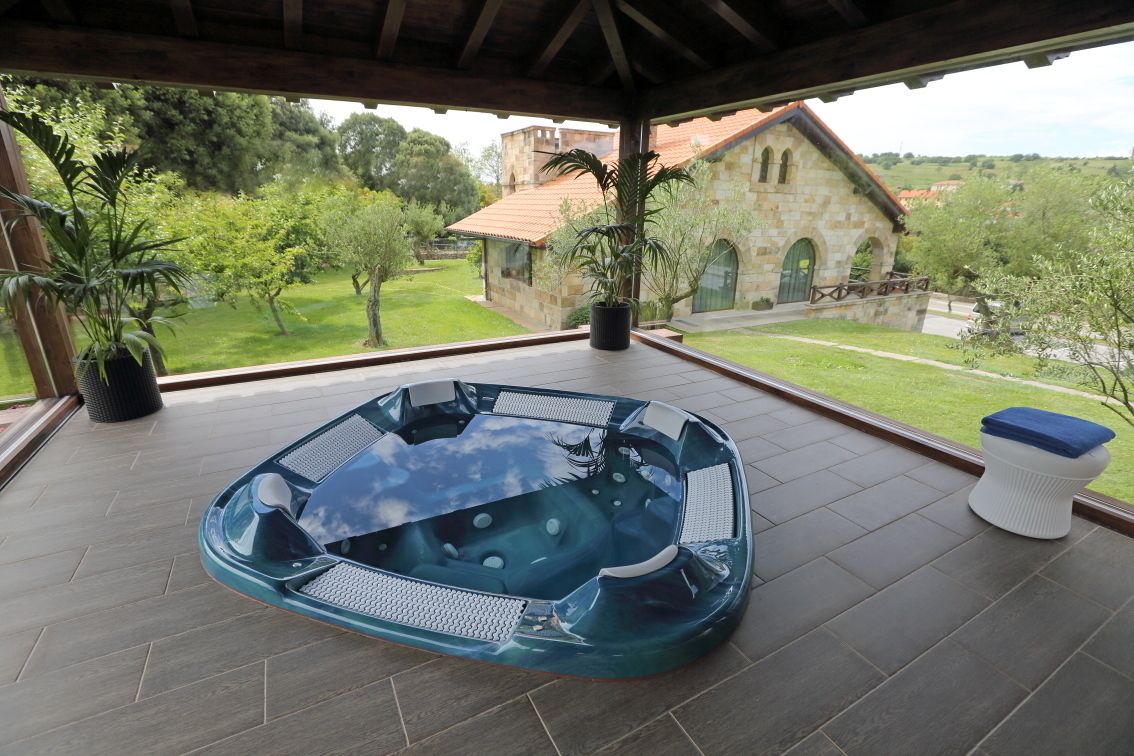 Un bain à remous sur une terrasse couverte, avec vue sur un bâtiment en pierre et une végétation luxuriante.