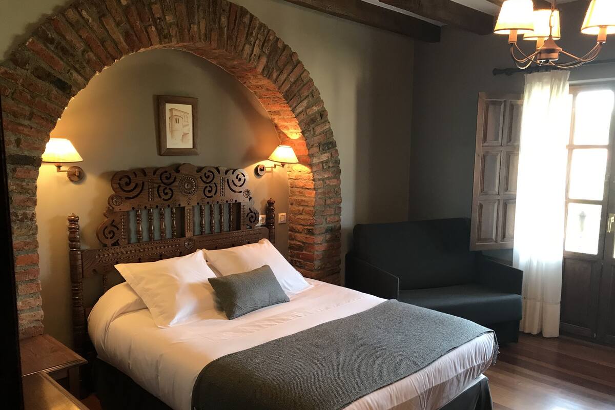 Uma cama com cabeceira de madeira, uma poltrona escura e um arco de tijolos.