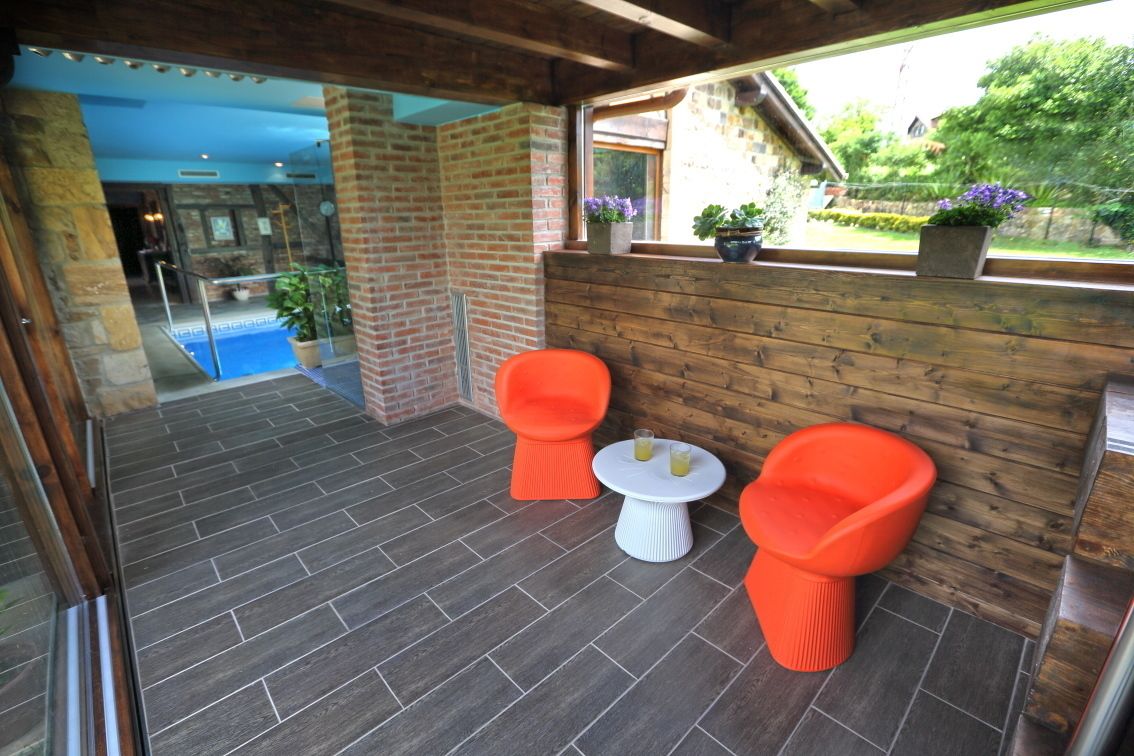 Un patio techado con pared de ladrillo, dos sillas naranjas, una mesita blanca y una ventana con plantas en macetas.