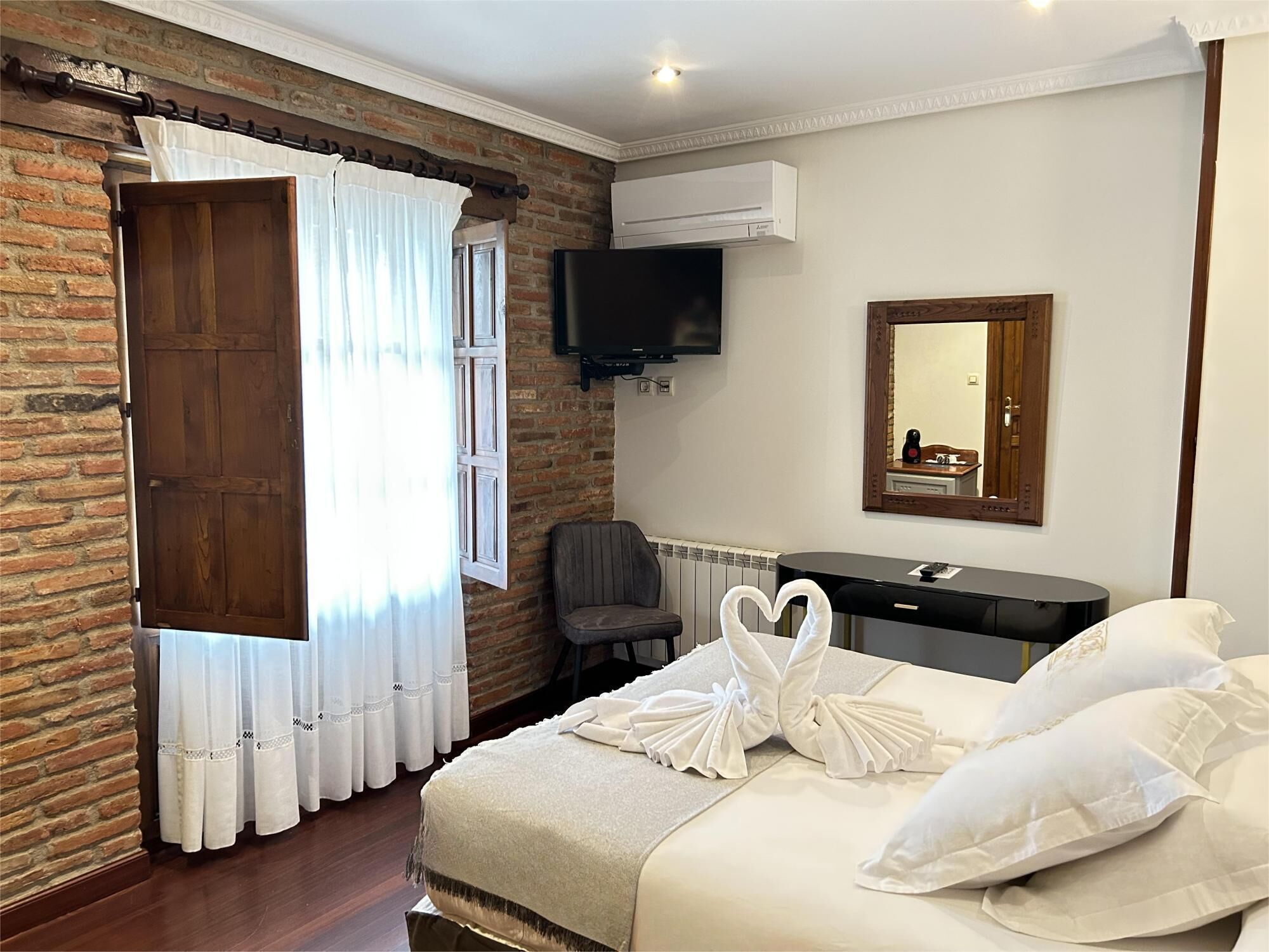 Ein Hotelzimmer mit Backsteinwand, Holztür, an der Wand befestigtem Fernseher, Spiegel, Schreibtisch und Bett mit Handtüchern, die als Herz angeordnet sind.