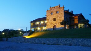 A large stone building with multiple balconies and a central tower, surrounded by a stone wall and a grassy slope. - Posada San Marcos (Santillana del Mar)