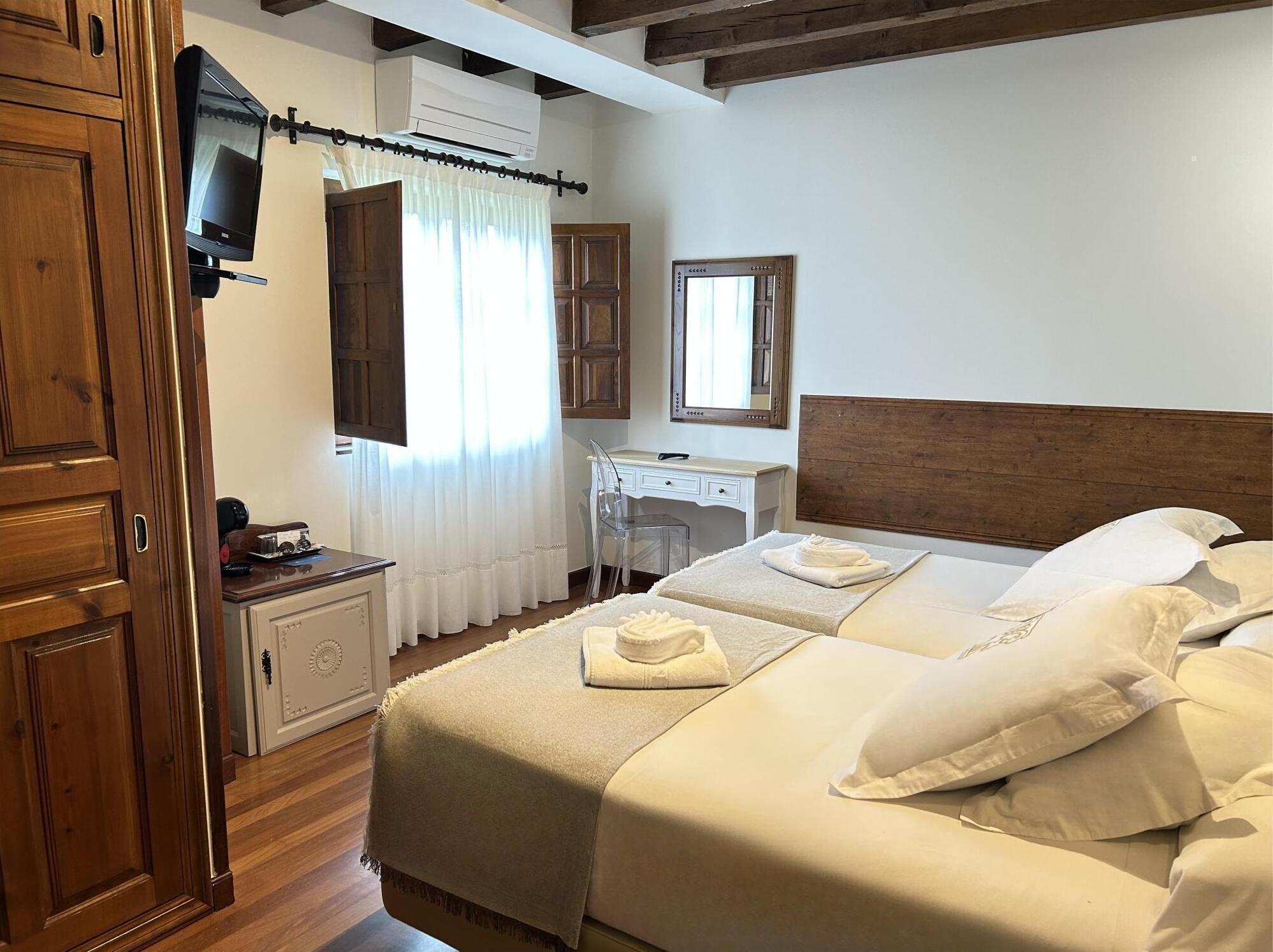 A hotel room with a wooden headboard, two beds with white linens, a desk with a chair, a mirror, and a television mounted on the wall.