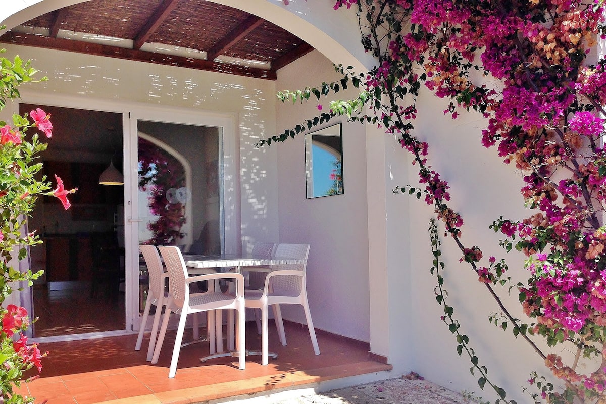 An outdoor patio with a table and chairs, a covered area, and a wall with flowering vines.