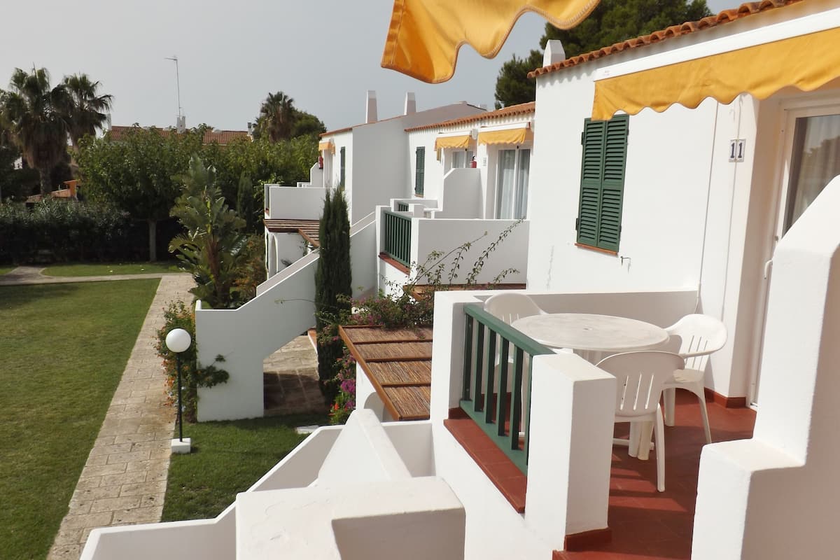 A white building with a balcony, a small garden, and a table with chairs.