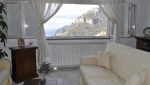 A living room with a white sofa, a wooden cabinet, and a view of a mountainous landscape through sheer curtains. - Bed & Breakfast Mamma Rosa (Positano)