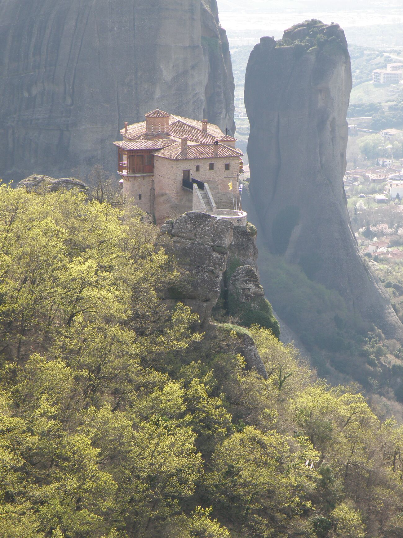 A stone building with a red-tiled roof perched on a cliff, surrounded by lush greenery and overlooking a valley.