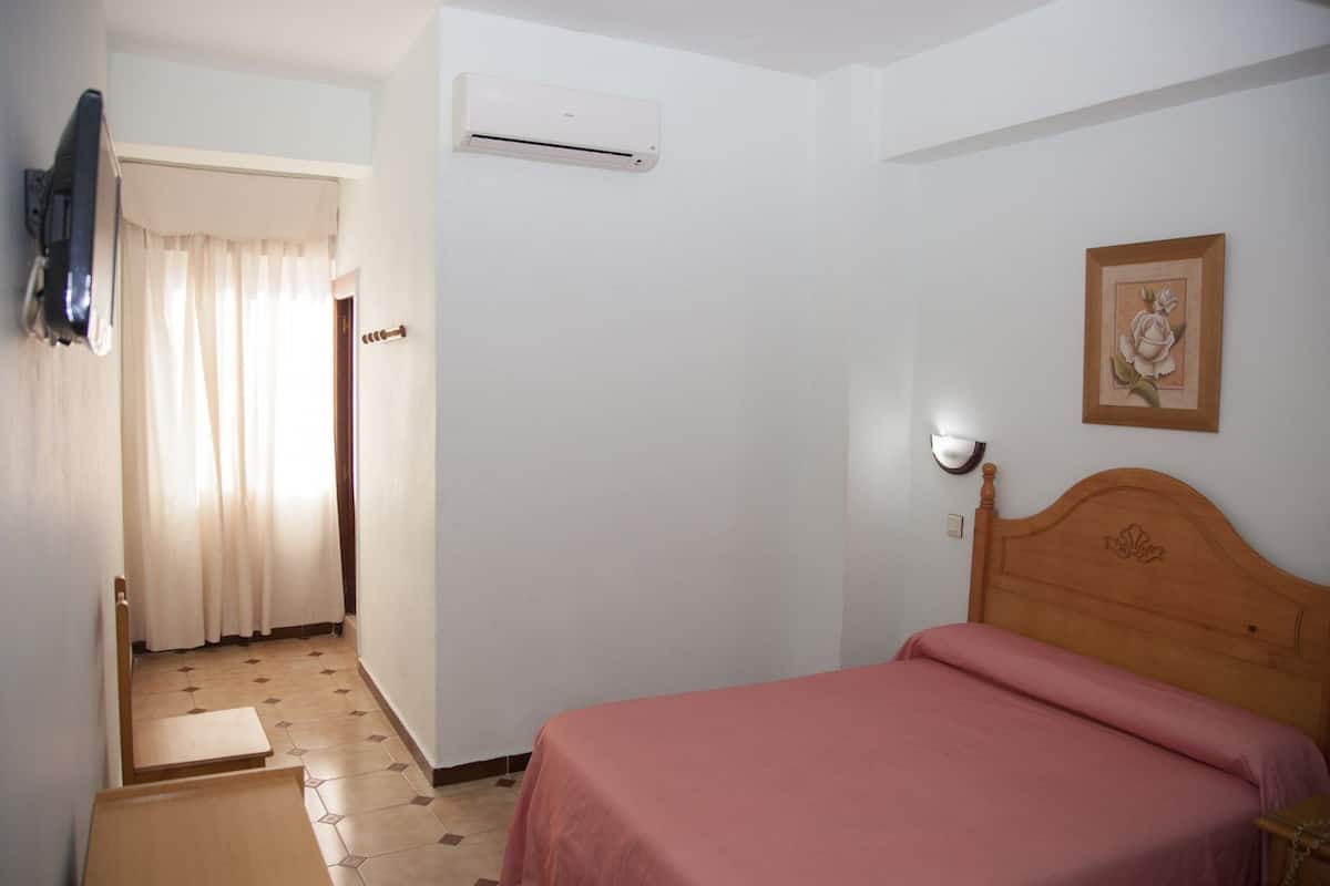 A bedroom with a bed, a bedside table, a wall-mounted air conditioner, a door leading to another room, and a framed picture on the wall.