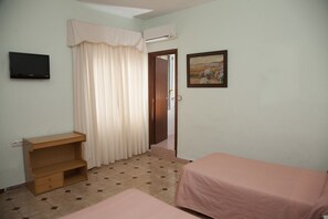 Una habitación pequeña y sencilla con una mesa de escritorio, dos camas, un cuadro y una puerta que da a otra habitación.
