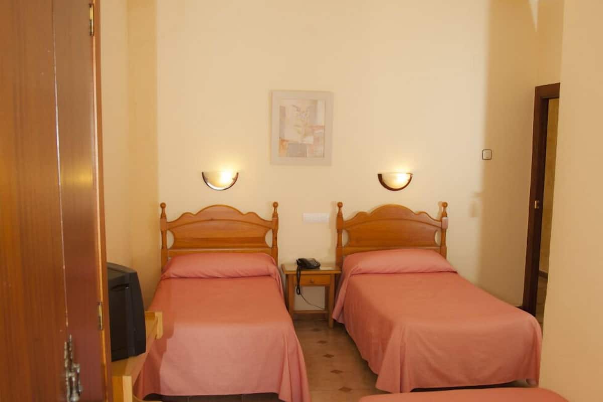 A room with two wooden beds, each with a headboard and a bedside table. There is a framed picture on the wall and a door on the left side.