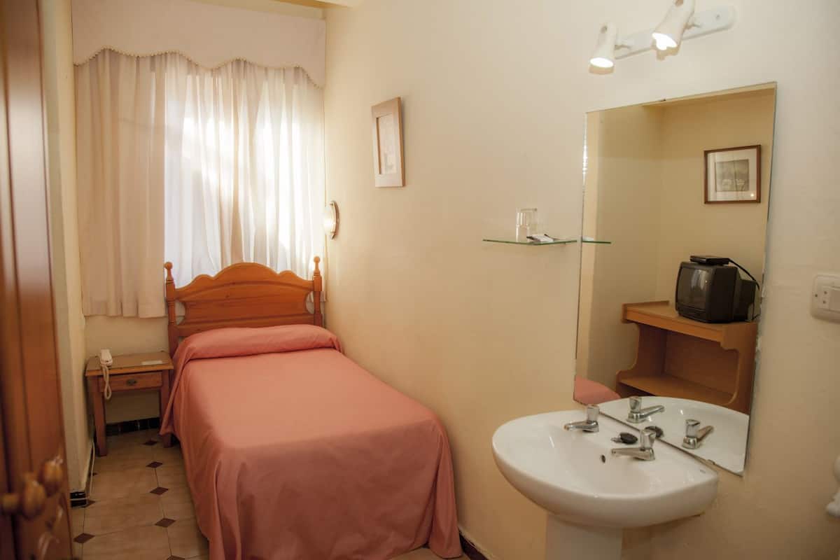 A bedroom with a single bed, a bedside table with a lamp, a mirror, a small shelf with a television, and a bathroom with a sink and mirror.