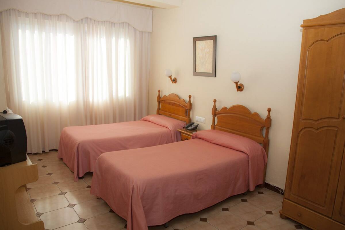 A hotel room with two single beds, a wooden wardrobe, a bedside table with a lamp, a telephone, and a framed picture on the wall.