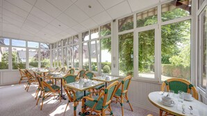 Een zonnige kamer met grote ramen, rieten stoelen en tafels gedekt voor het eten.