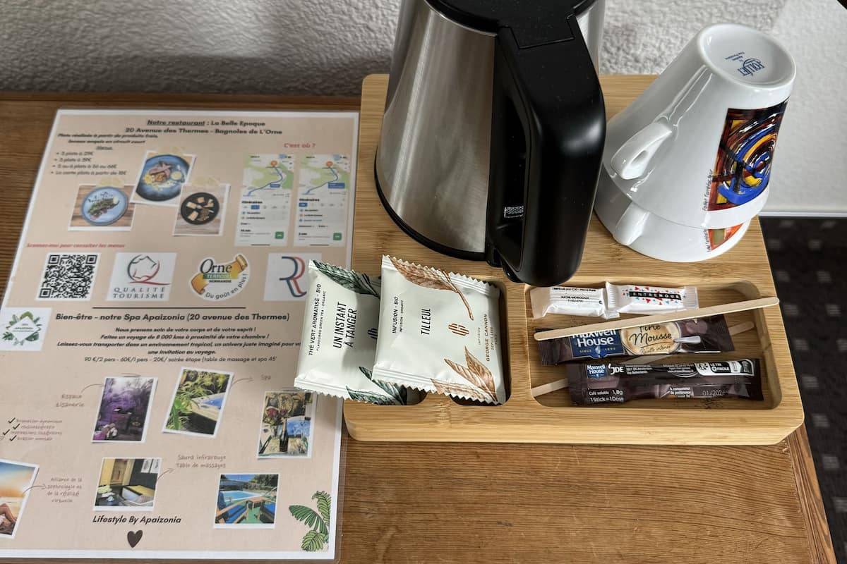 Een houten tafel met een roestvrijstalen waterkoker, een witte mok met een kleurrijk ontwerp, en een dienblad met diverse spullen, waaronder een brochure, een QR-code en wat snacks.