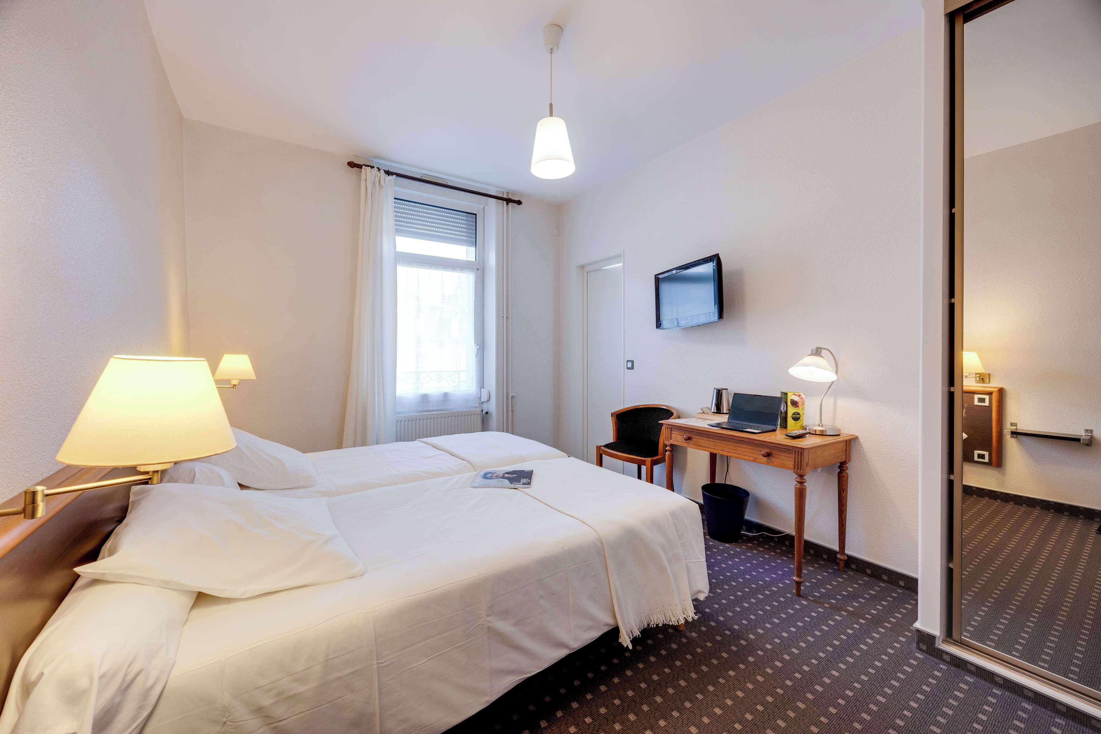 Une chambre d’hôtel avec deux lits, un bureau avec un ordinateur, une chaise, un téléviseur fixé au mur et une porte avec un miroir.