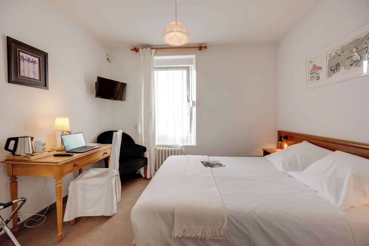 Een hotelkamer met een bed, een bureau met een laptop, een stoel, een televisie en een raam met gordijnen.