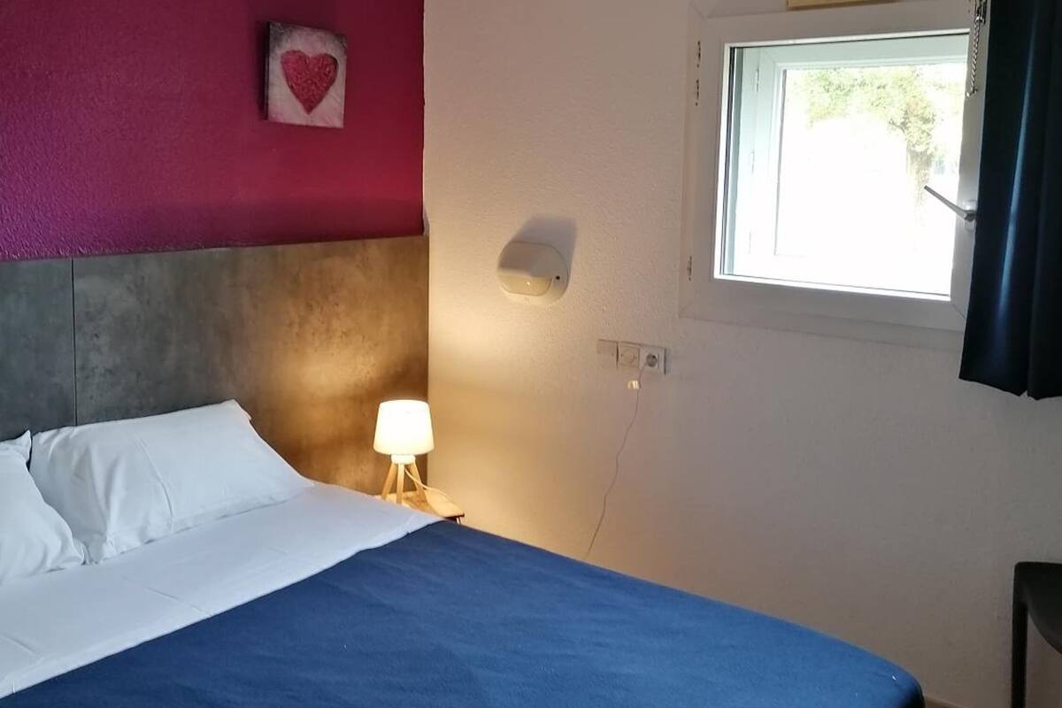 A bedroom with a bed, a bedside lamp, a window, and a wall-mounted light fixture.