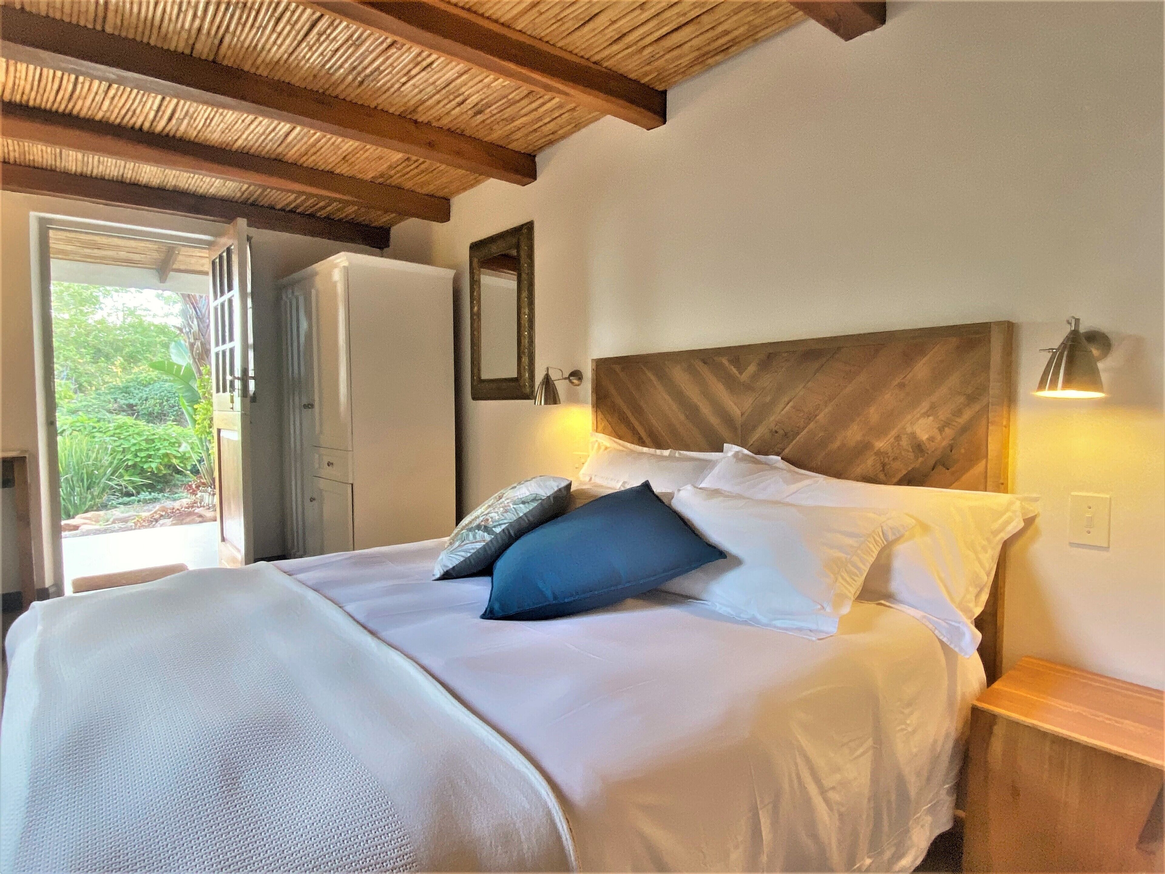 A bedroom with a bed, a wooden headboard, a nightstand with a lamp, a mirror, and a door leading to a garden.