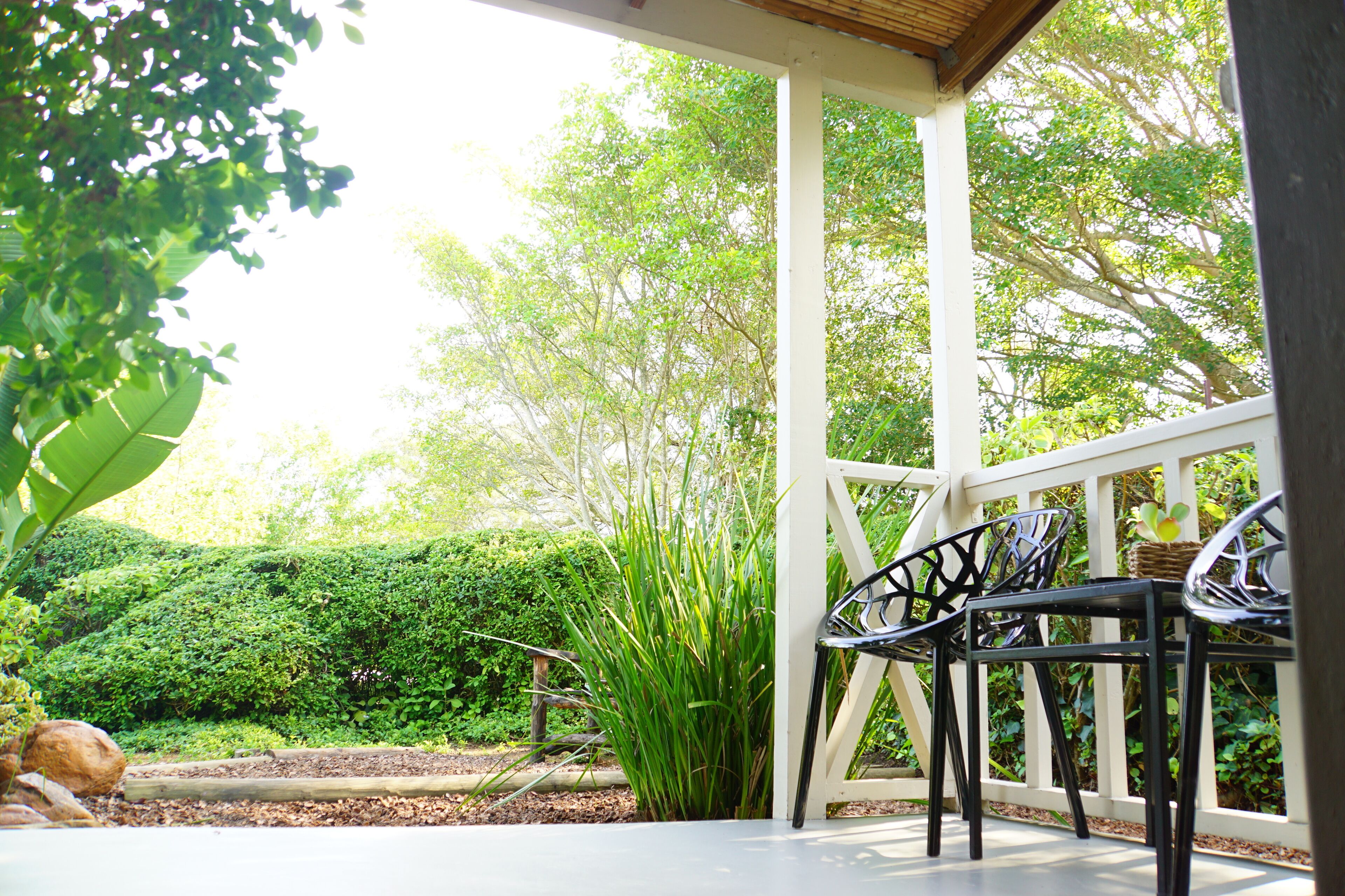 A covered outdoor area with a white railing, two black metal chairs, and lush greenery in the background.