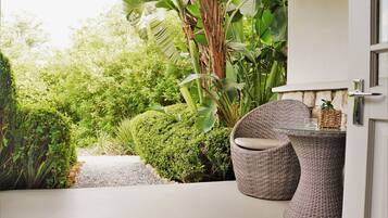 Relax in a serene outdoor seating area with a wicker chair and glass-top table overlooking a lush garden.