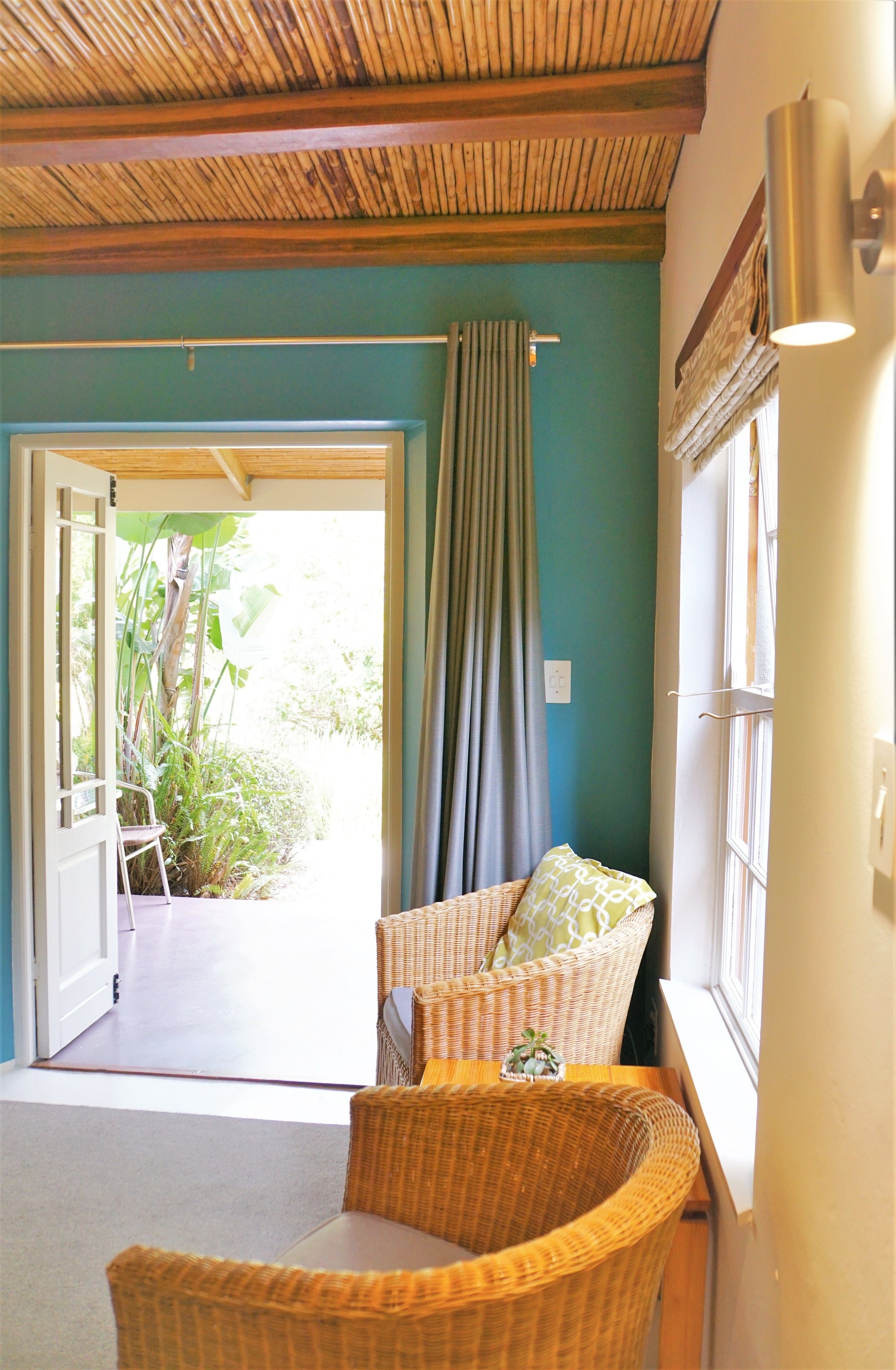 A room with a wicker chair, a small table, a window with a curtain, and a door leading to an outdoor area with plants.