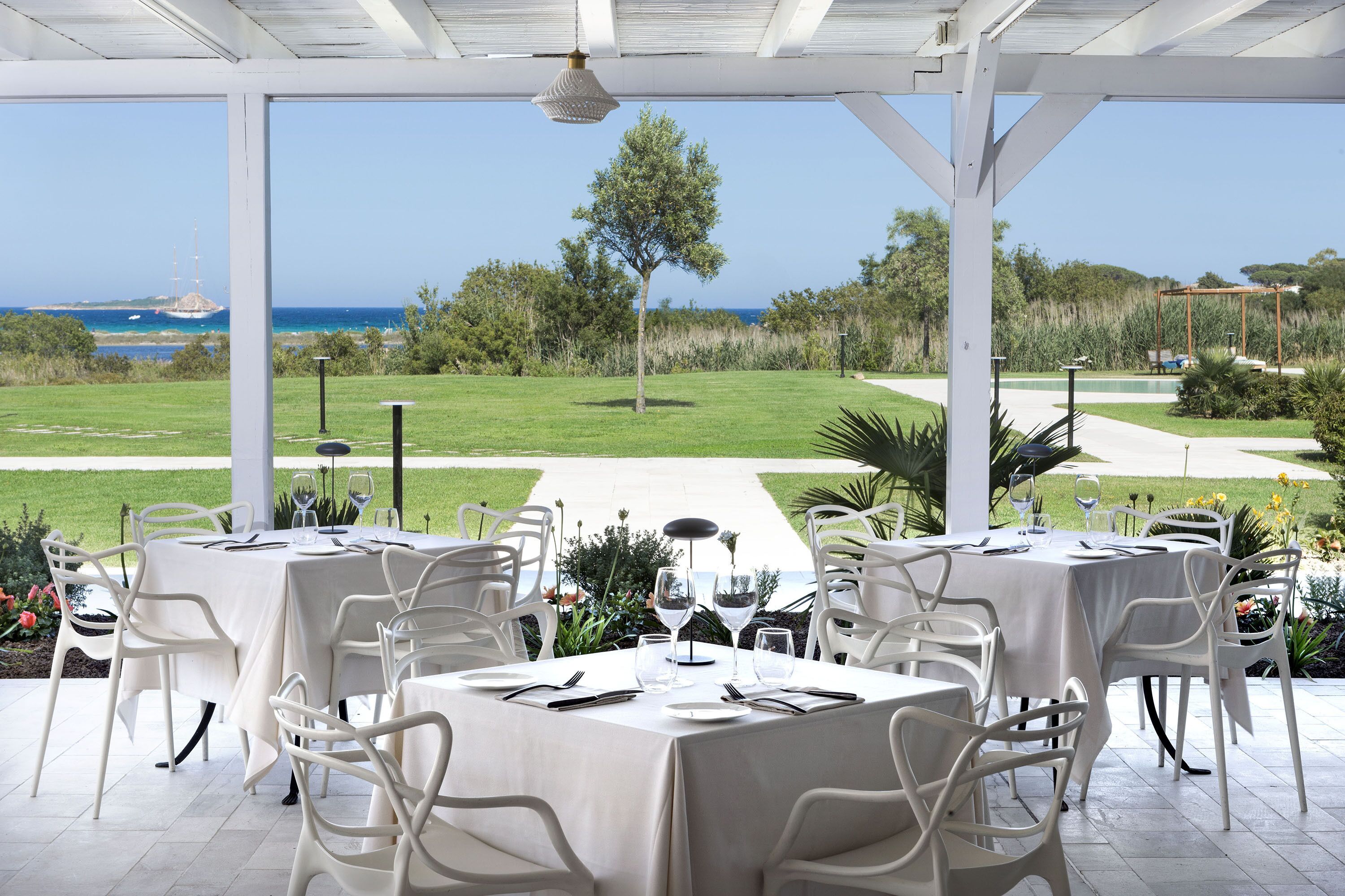 Un espacio de comedor al aire libre con mesas y sillas blancas, con vistas a un campo de golf y al océano.
