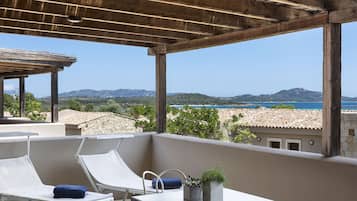 A balcony with two white lounge chairs, a small table, and a potted plant. The balcony overlooks a scenic view of the sea and distant hills.