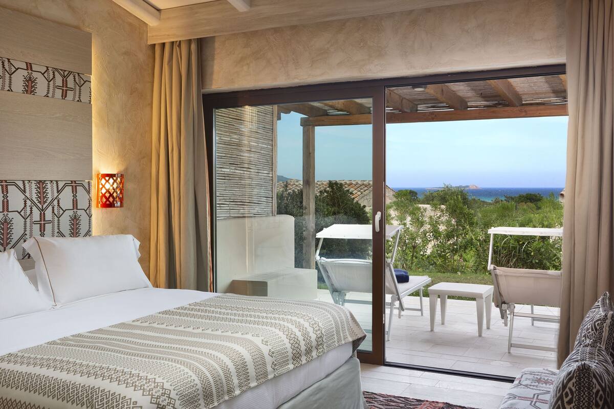 Habitación de hotel con cama, una mesita y una silla. Posee una puerta corrediza de vidrio que da a un espacio exterior con vistas al océano.