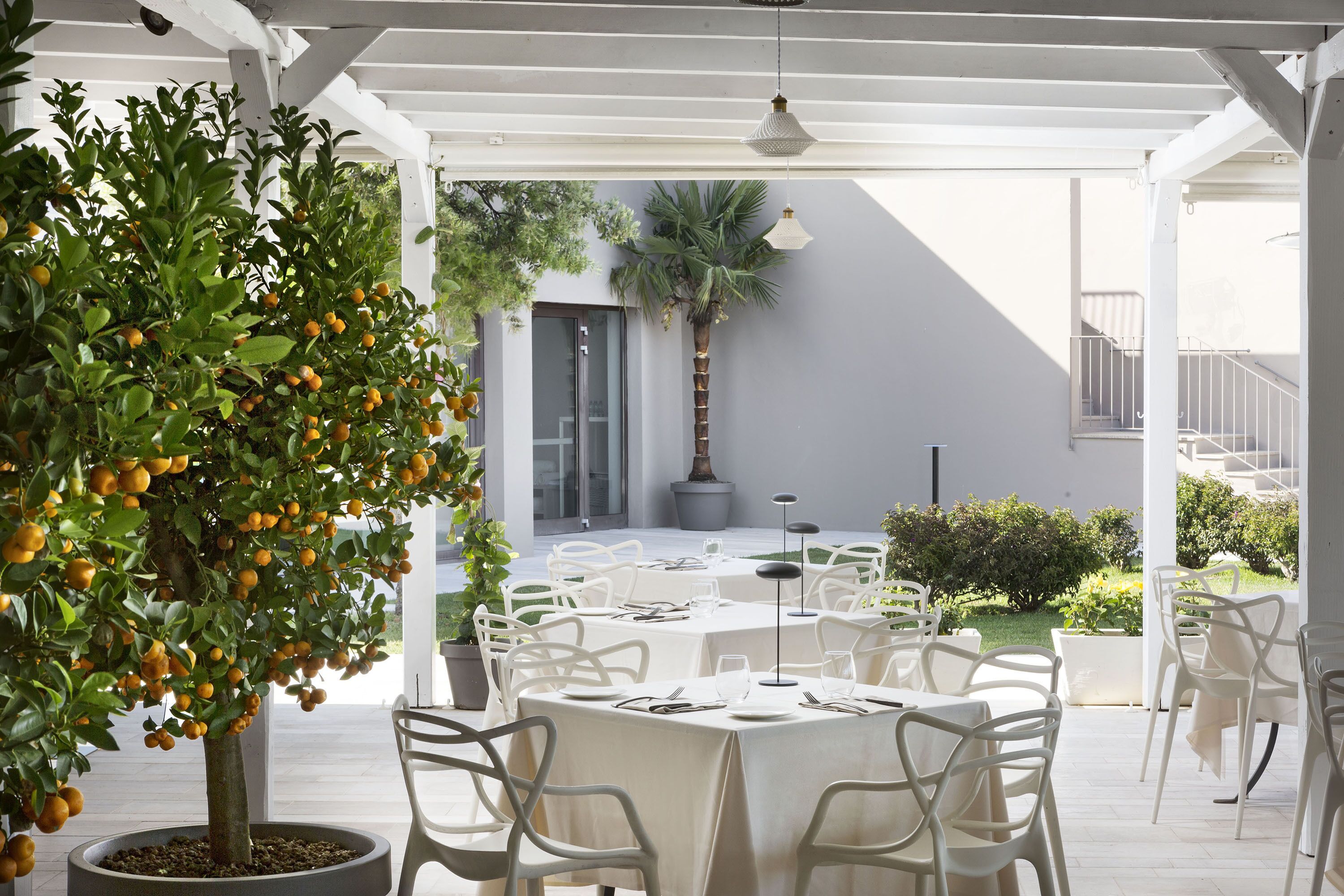 An outdoor dining area with white tables and chairs, a potted lemon tree, and a staircase leading to an upper level.