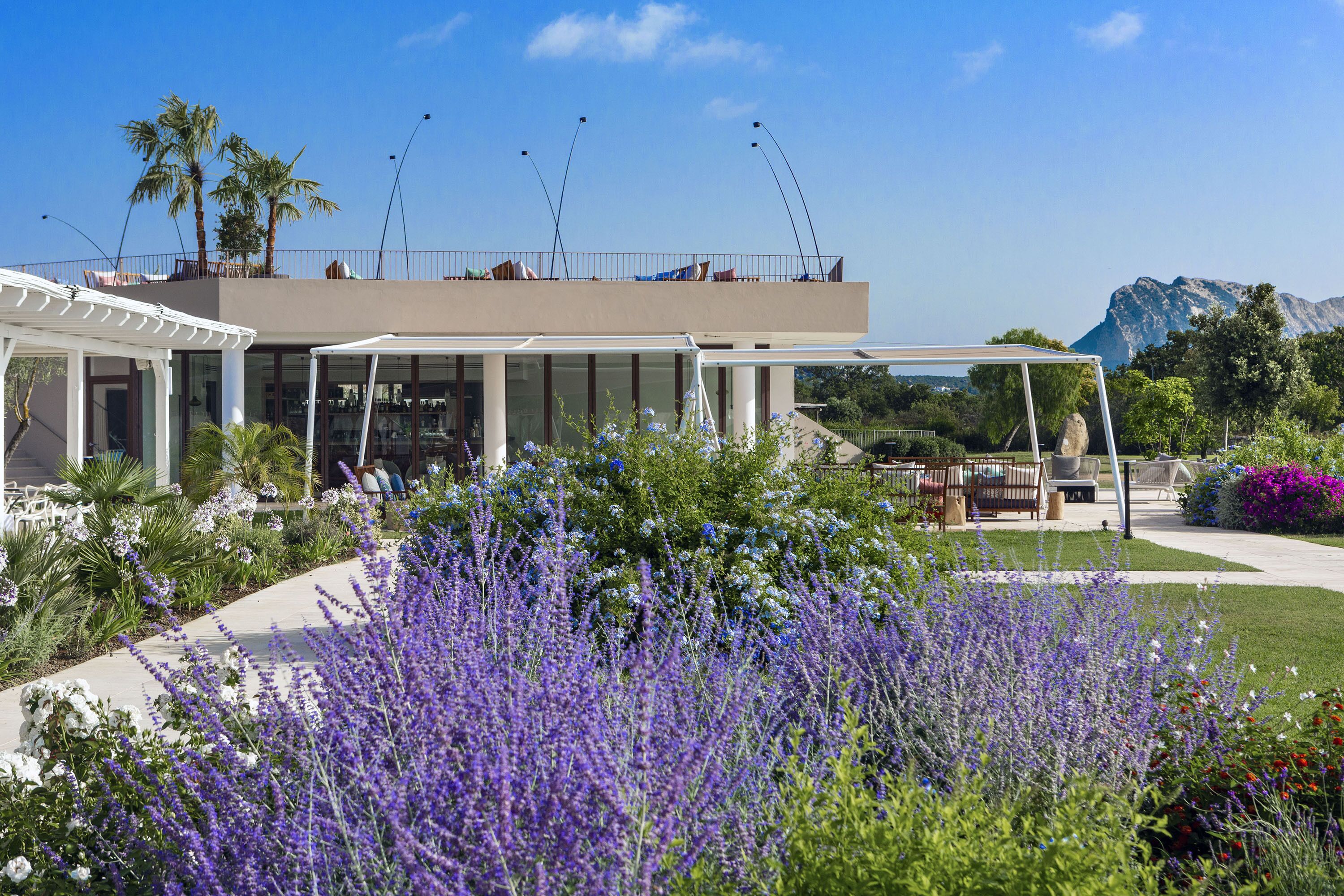 A modern building with a covered patio area, surrounded by lush gardens and a mountain backdrop.