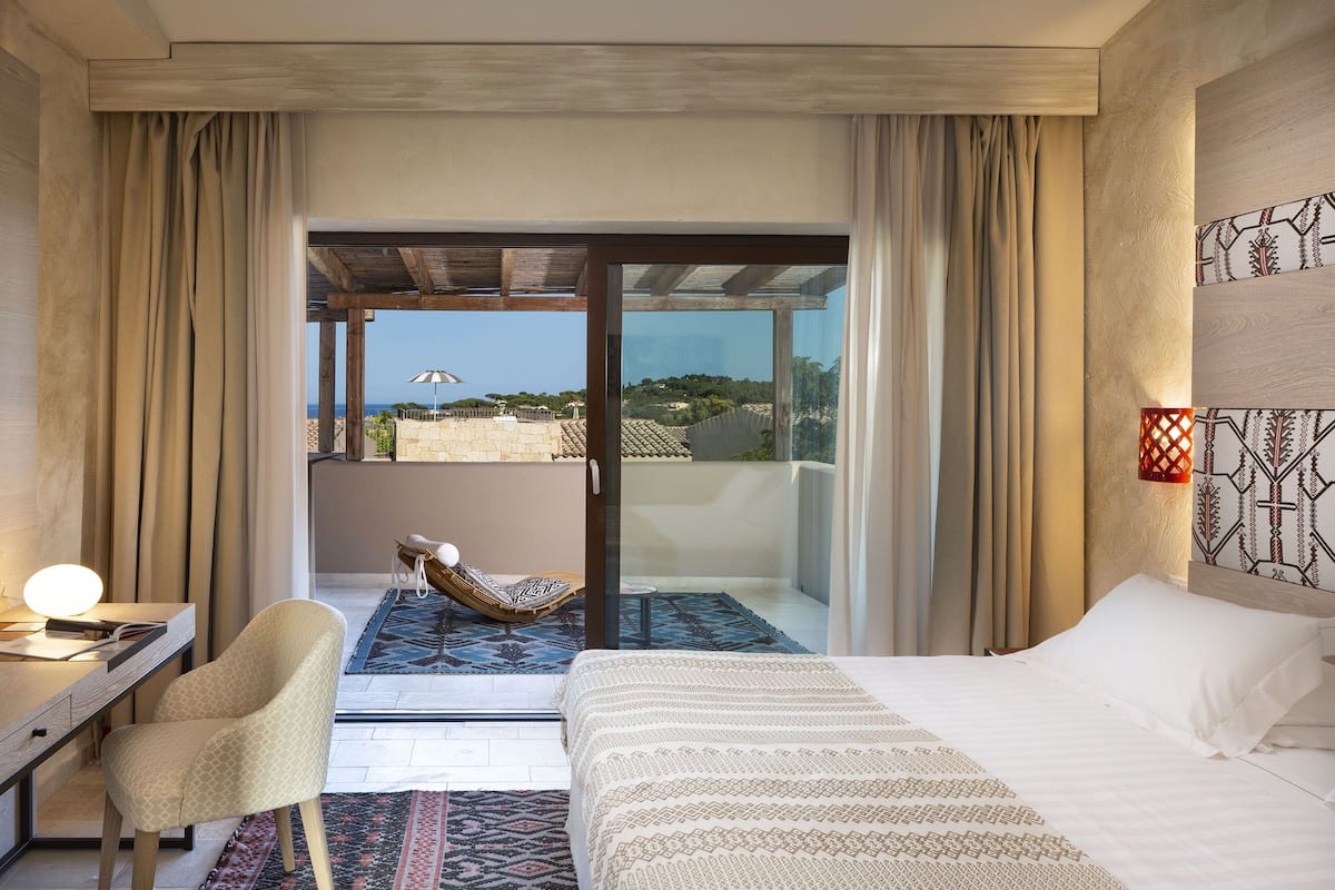 Habitación de hotel con cama, escritorio con lámpara, silla, alfombra estampada y vista al mar a través de una puerta de vidrio.