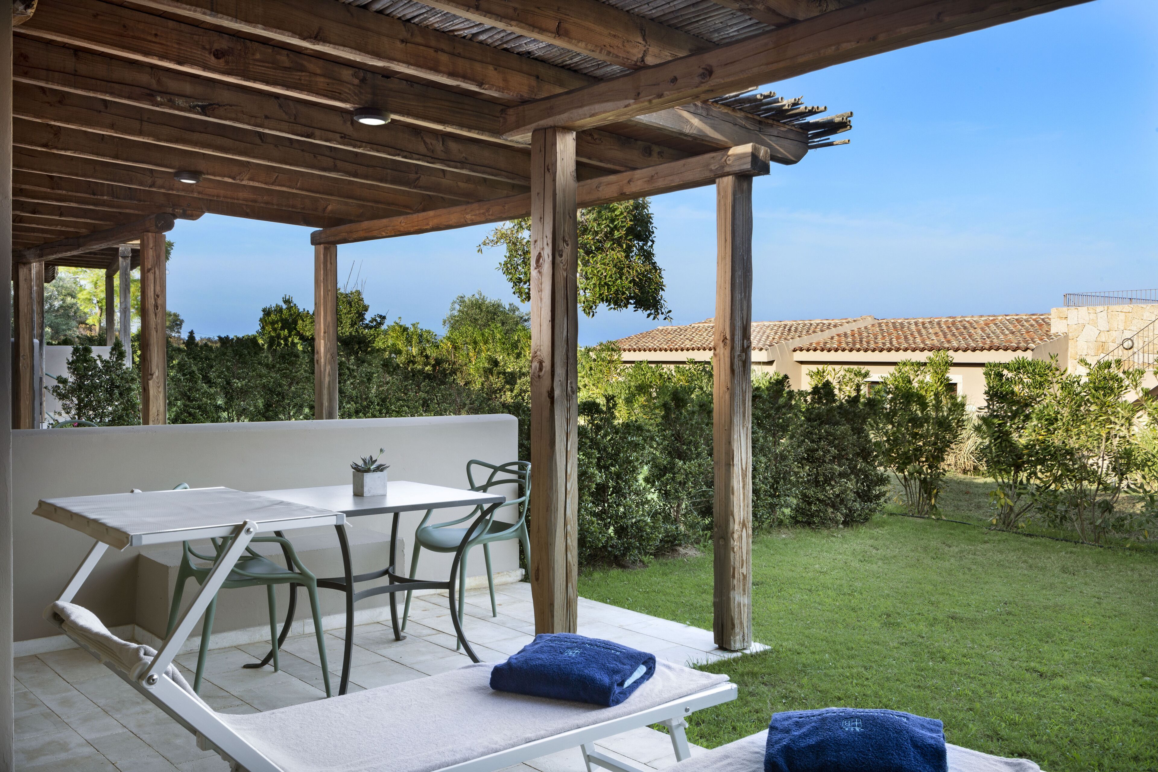 Un área de estar al aire libre techada, con una mesa y sillas, una planta pequeña en maceta y vistas a un jardín y edificios al fondo.
