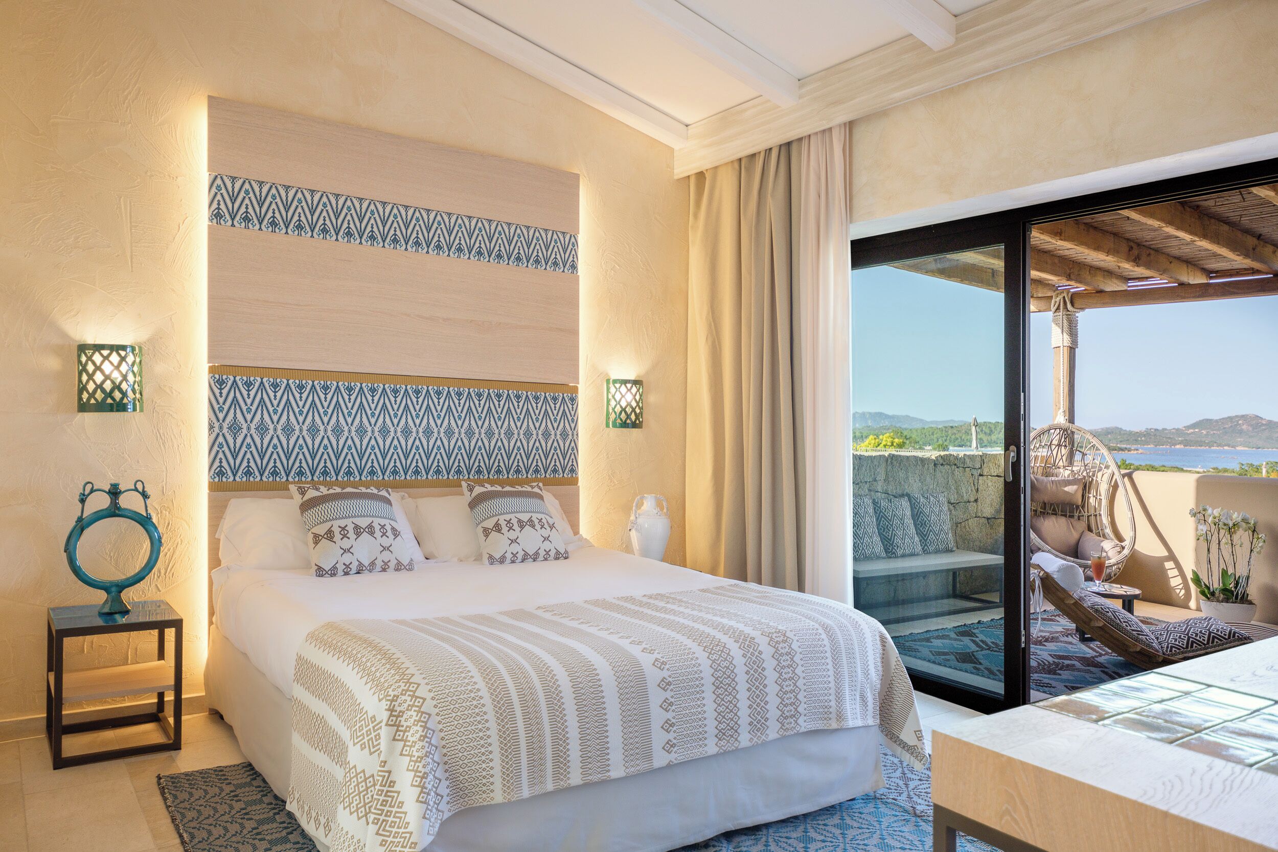 A bedroom with a bed, bedside lamps, a nightstand, a view of the outdoors through a sliding door, and a balcony with a chair.
