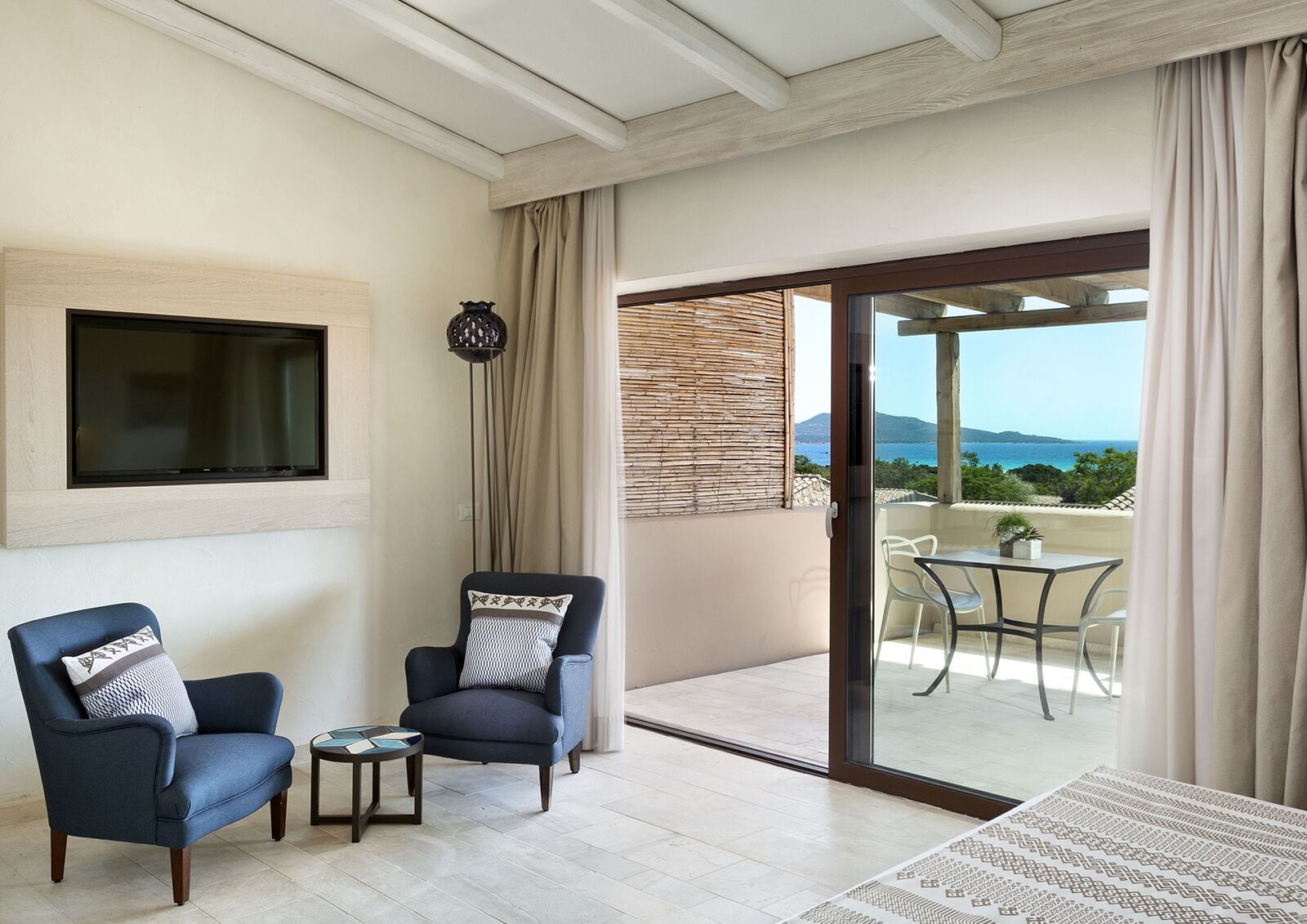 A living room with two armchairs, a small table, and a TV mounted on the wall. There is a sliding glass door leading to an outdoor area with a table and chairs.