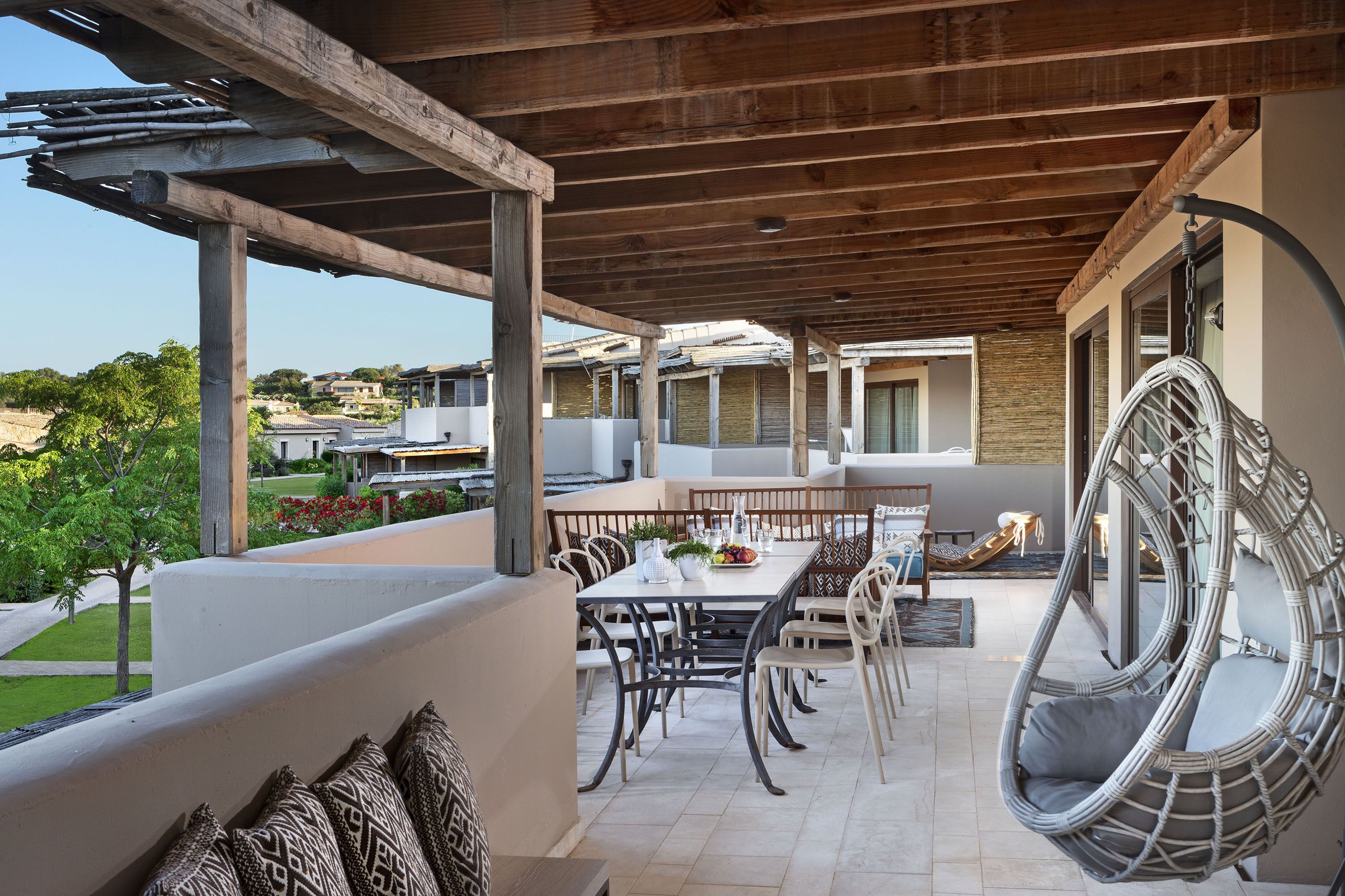 A covered outdoor dining area with a table and chairs, a hanging chair, and a view of the surrounding landscape.