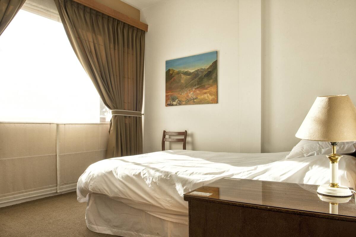 Una habitación de hotel con cama, mesita de noche, lámpara, un cuadro en la pared y ventana con cortinas.