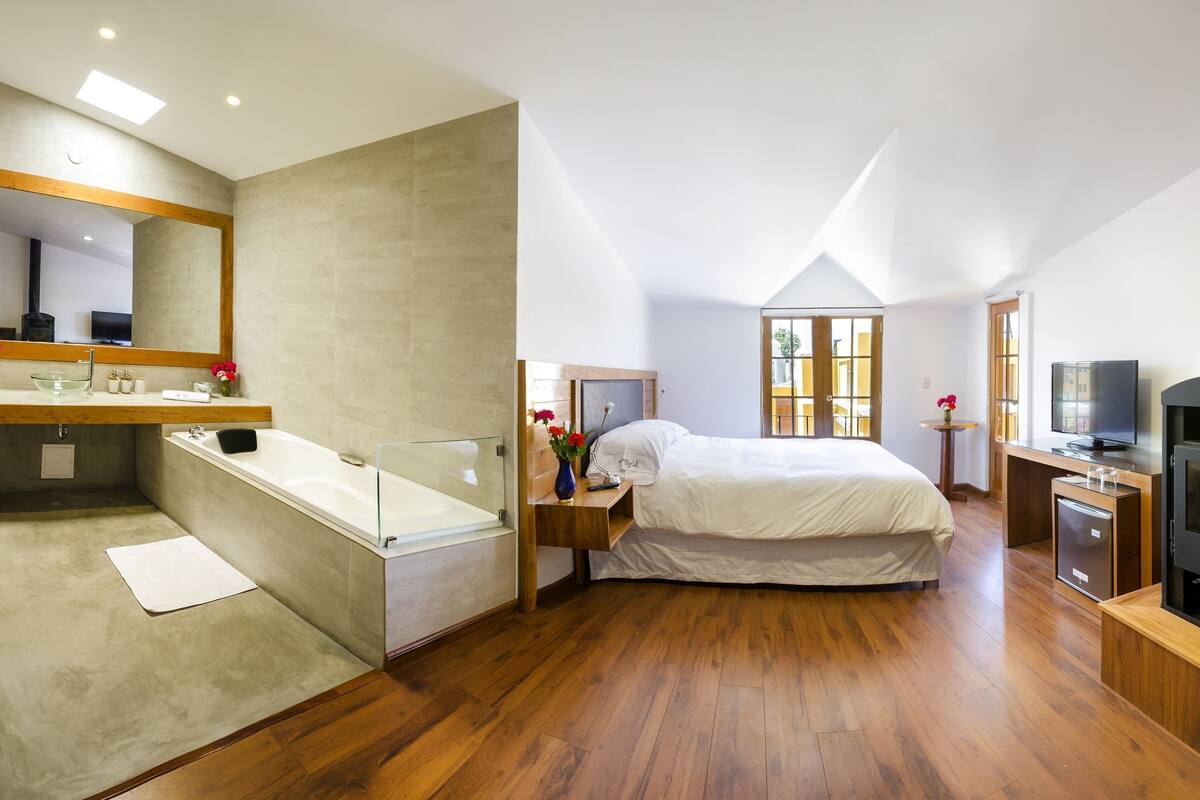 Un dormitorio con una cama grande, piso de madera, bañera de vidrio, televisor y chimenea.