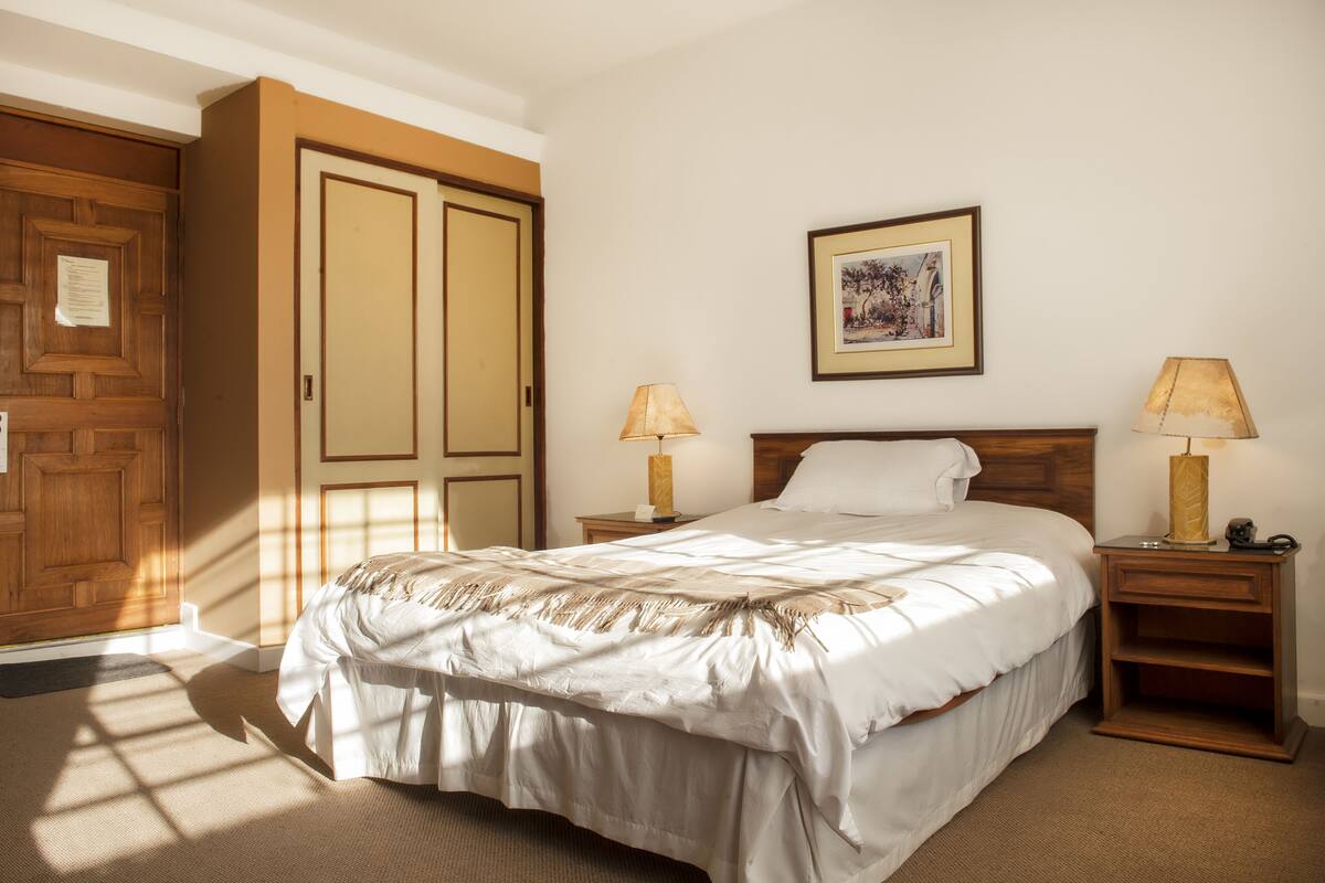 Un dormitorio con una cama, dos mesitas de noche con lámparas, un armario de madera y un cuadro enmarcado en la pared.