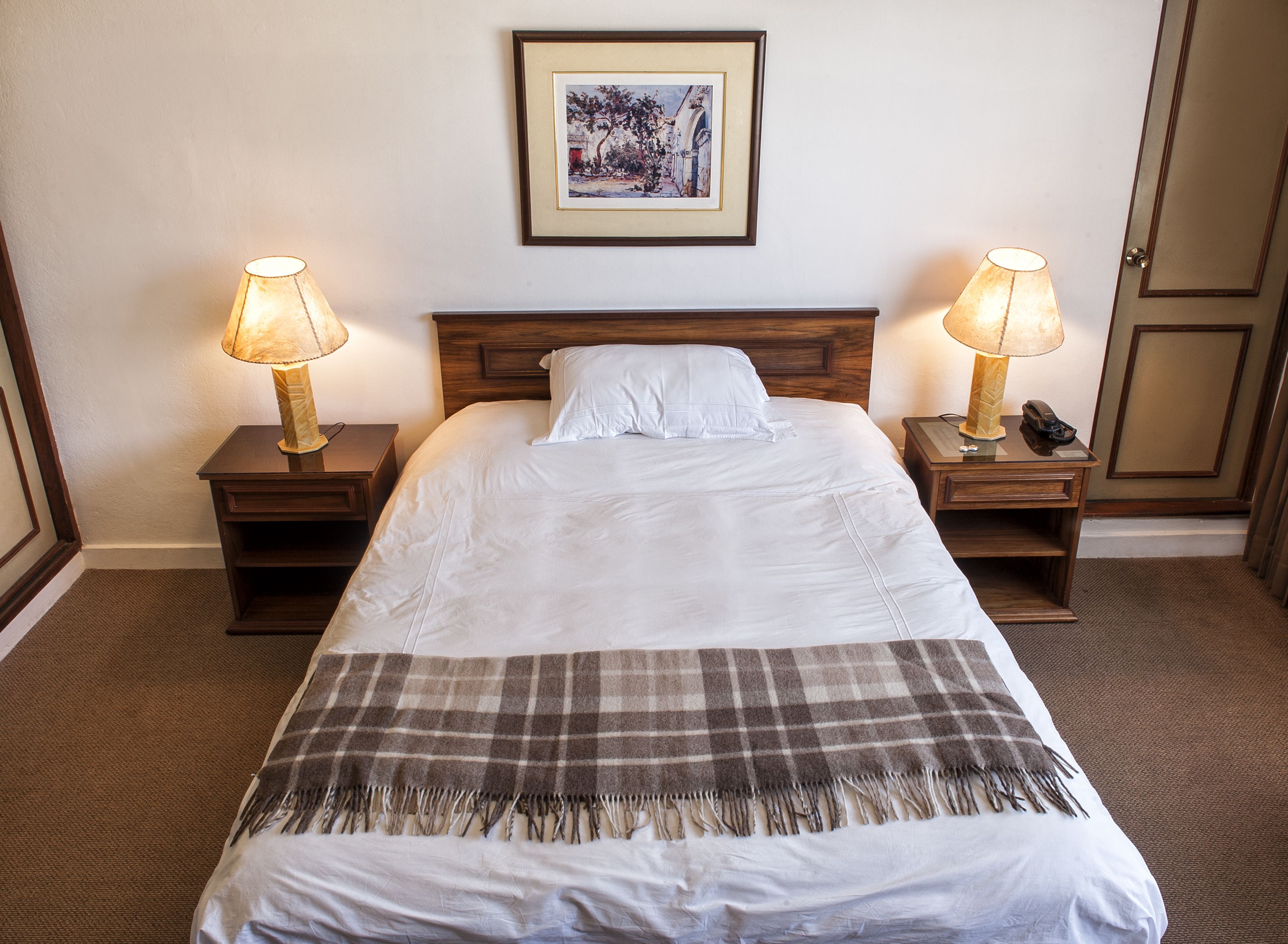 A neatly made bed with a plaid blanket, two bedside lamps, a framed picture on the wall, and a wooden headboard.